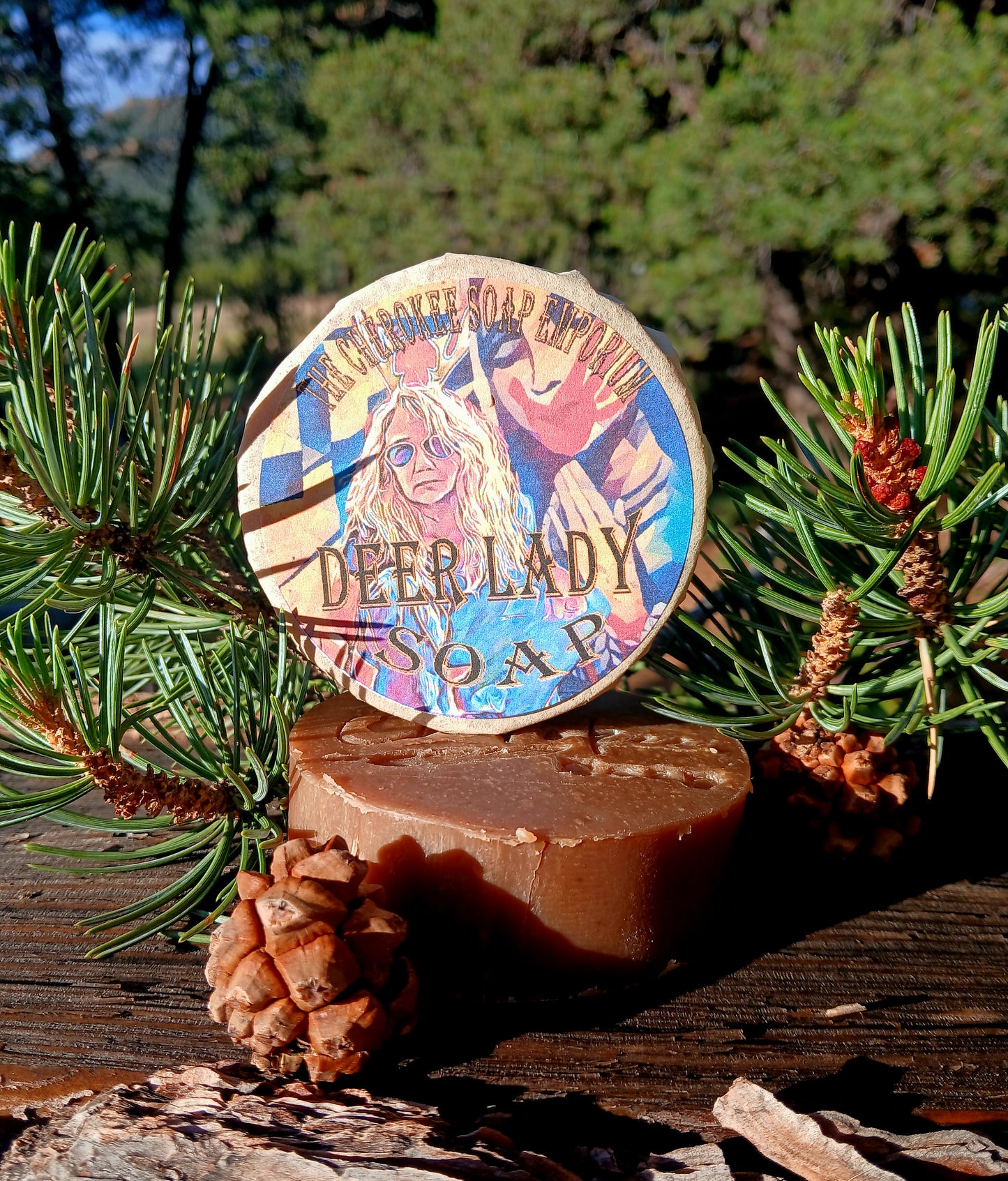 A round bar of Deer Lady Soap stands upright on a larger rectangular bar, photographed outdoors on a wooden surface with pine branches and pinecones around it. The top bar has a colorful illustrated label reading 'DEER LADY SOAP' and 'Misty Mountain Cream Soap.