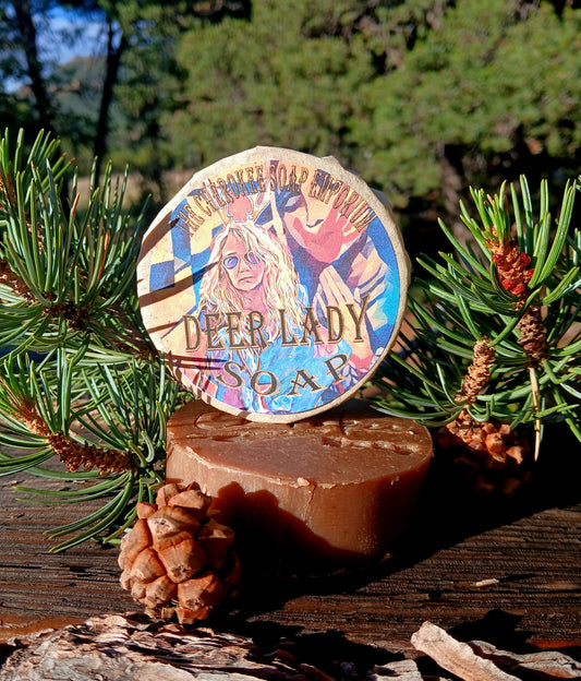 A round bar of Deer Lady Soap stands upright on a larger rectangular bar, photographed outdoors on a wooden surface with pine branches and pinecones around it. The top bar has a colorful illustrated label reading 'DEER LADY SOAP' and 'Misty Mountain Cream Soap.